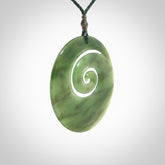 This picture shows a large hand-carved koru. It is carved from British Columbian jade and is suspended from a hand-plaited, green cord.